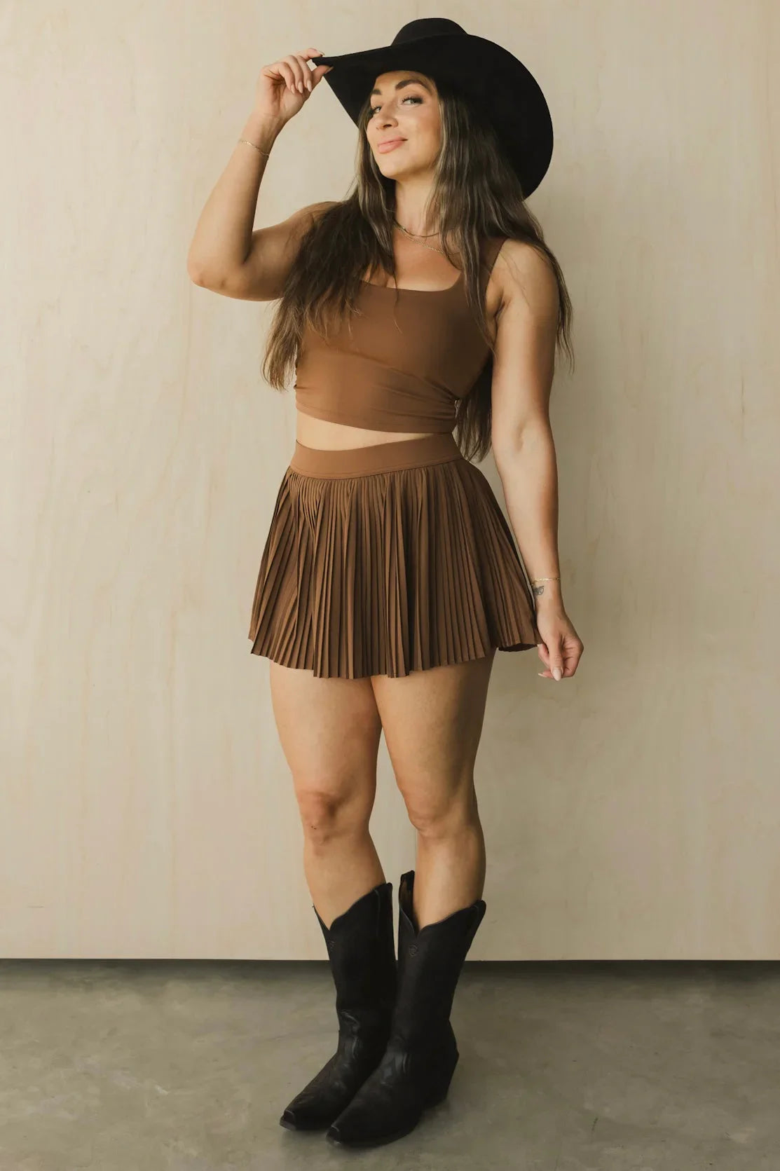 Woman in brown crop top and pleated mini skirt, black cowboy boots, and black hat