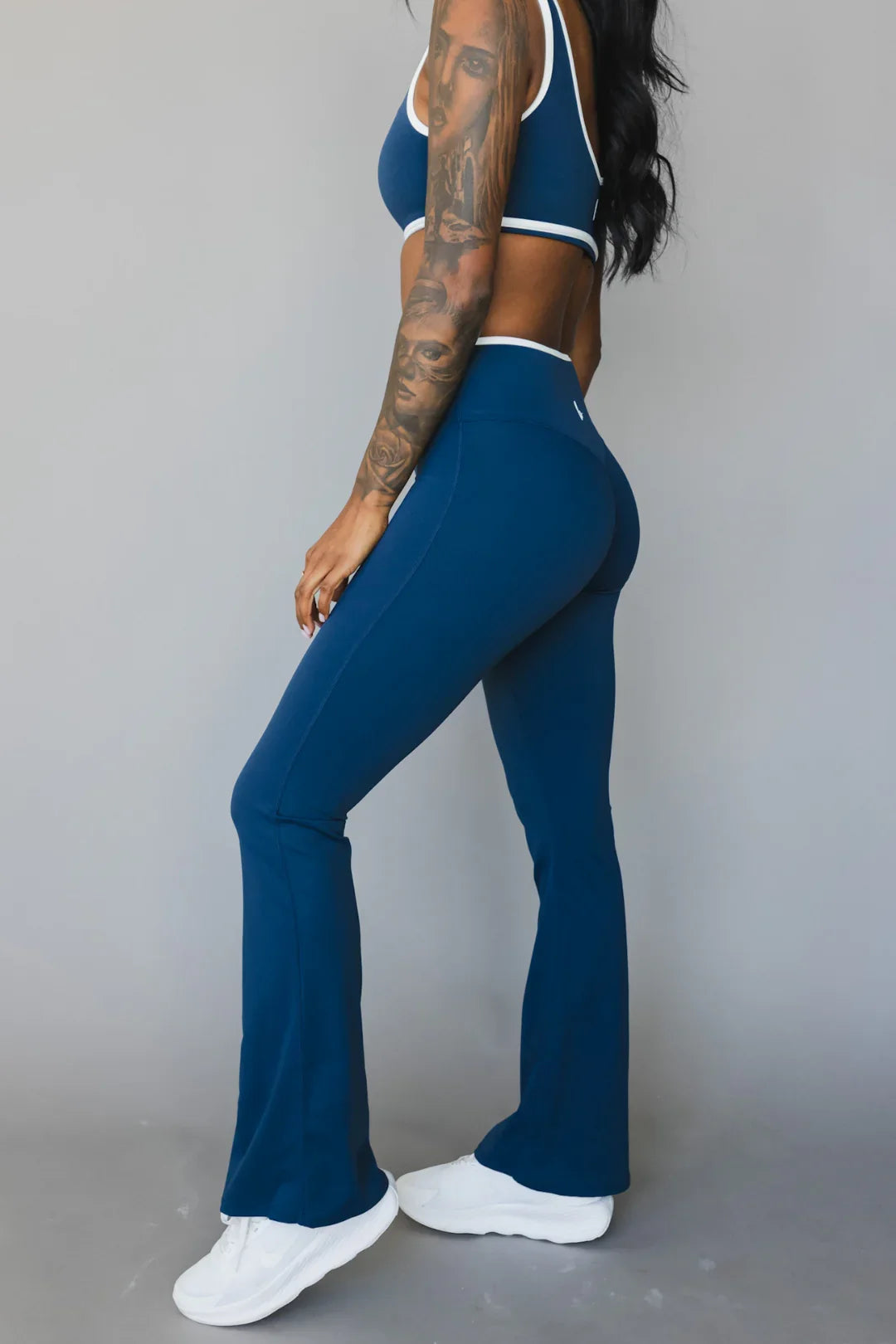 Woman in blue athletic flared leggings and sports bra, showcasing tattoos and white sneakers