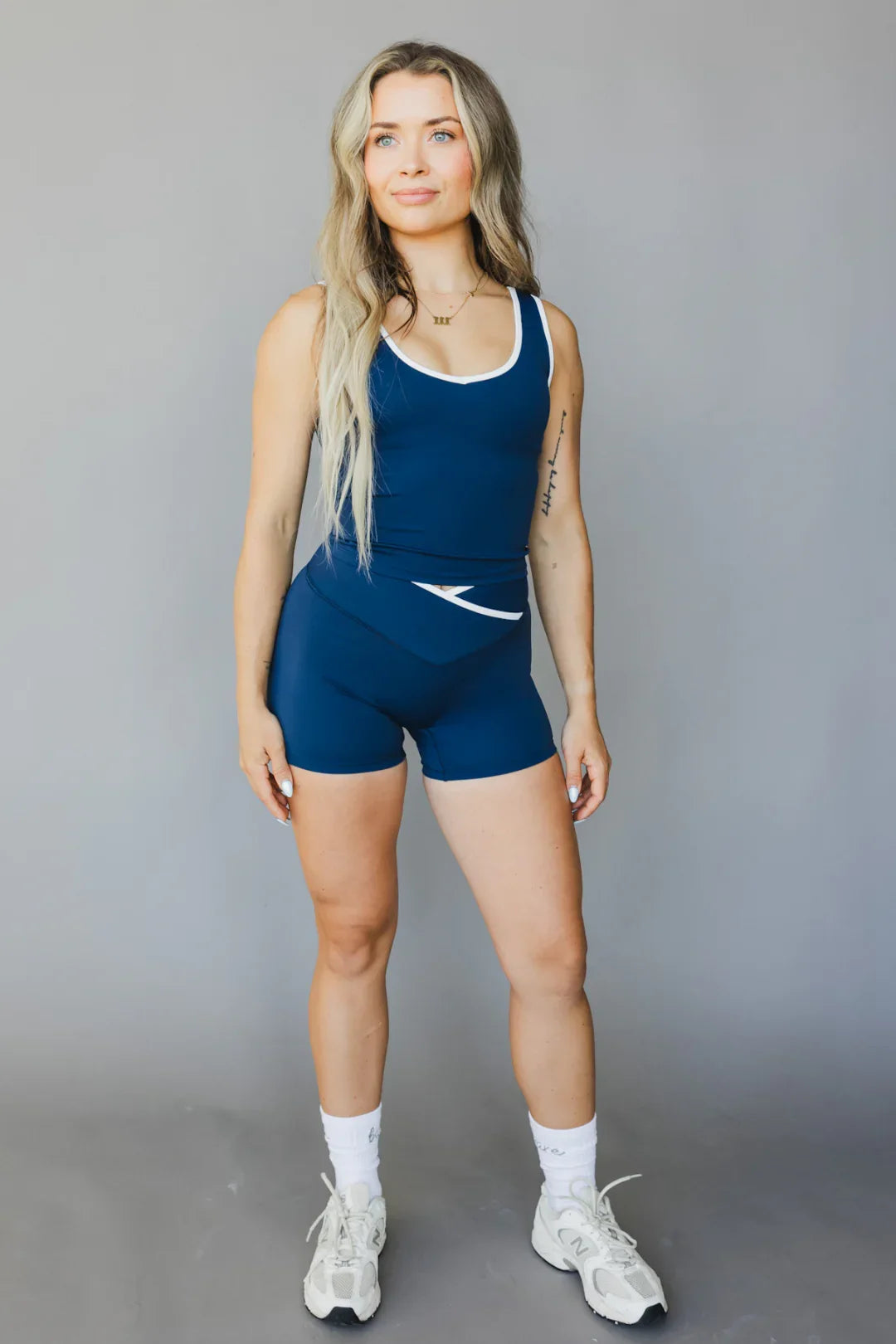 Woman wearing blue athletic tank top and shorts set, white sneakers, gray studio background