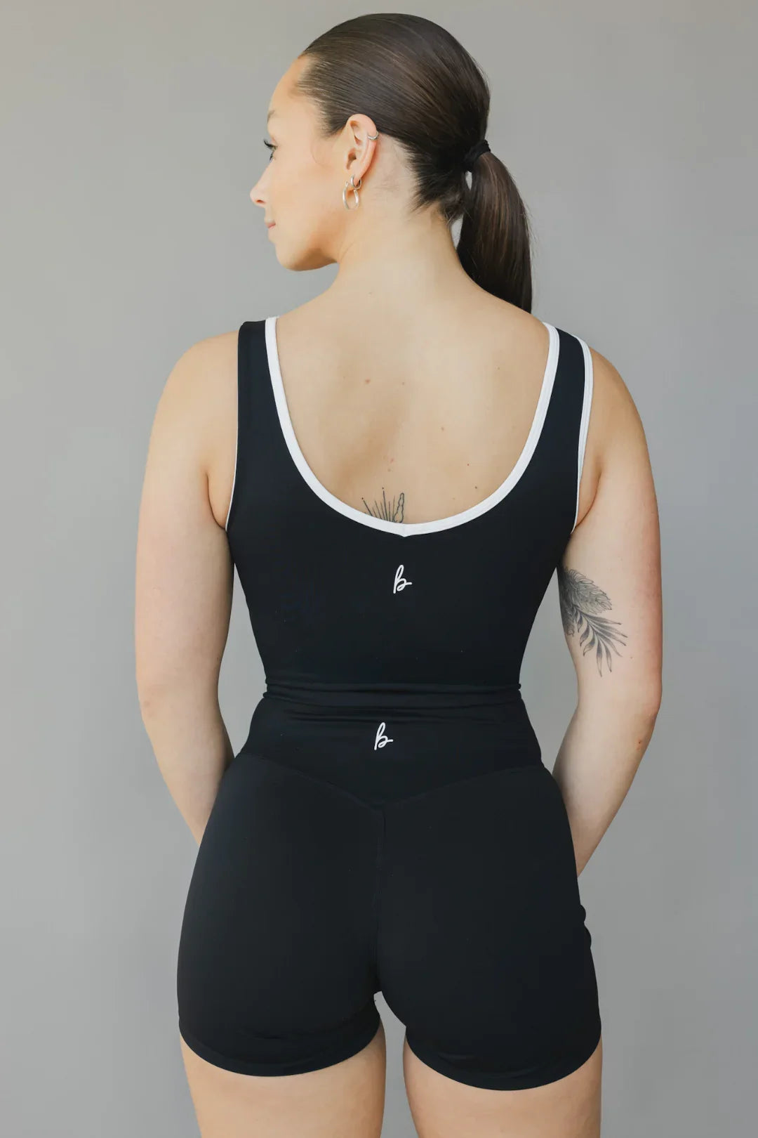 Woman in black athletic romper with white trim, back view, showing tattoos and ponytail
