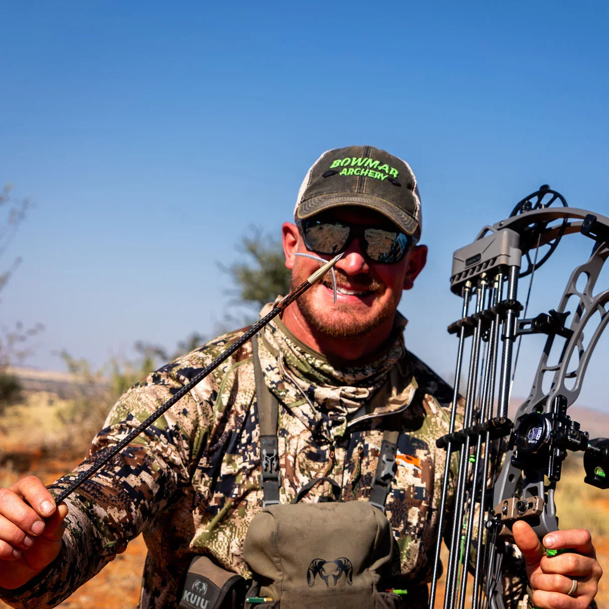 Hunter in camo outfit holding compound bow and arrow outdoors under blue sky