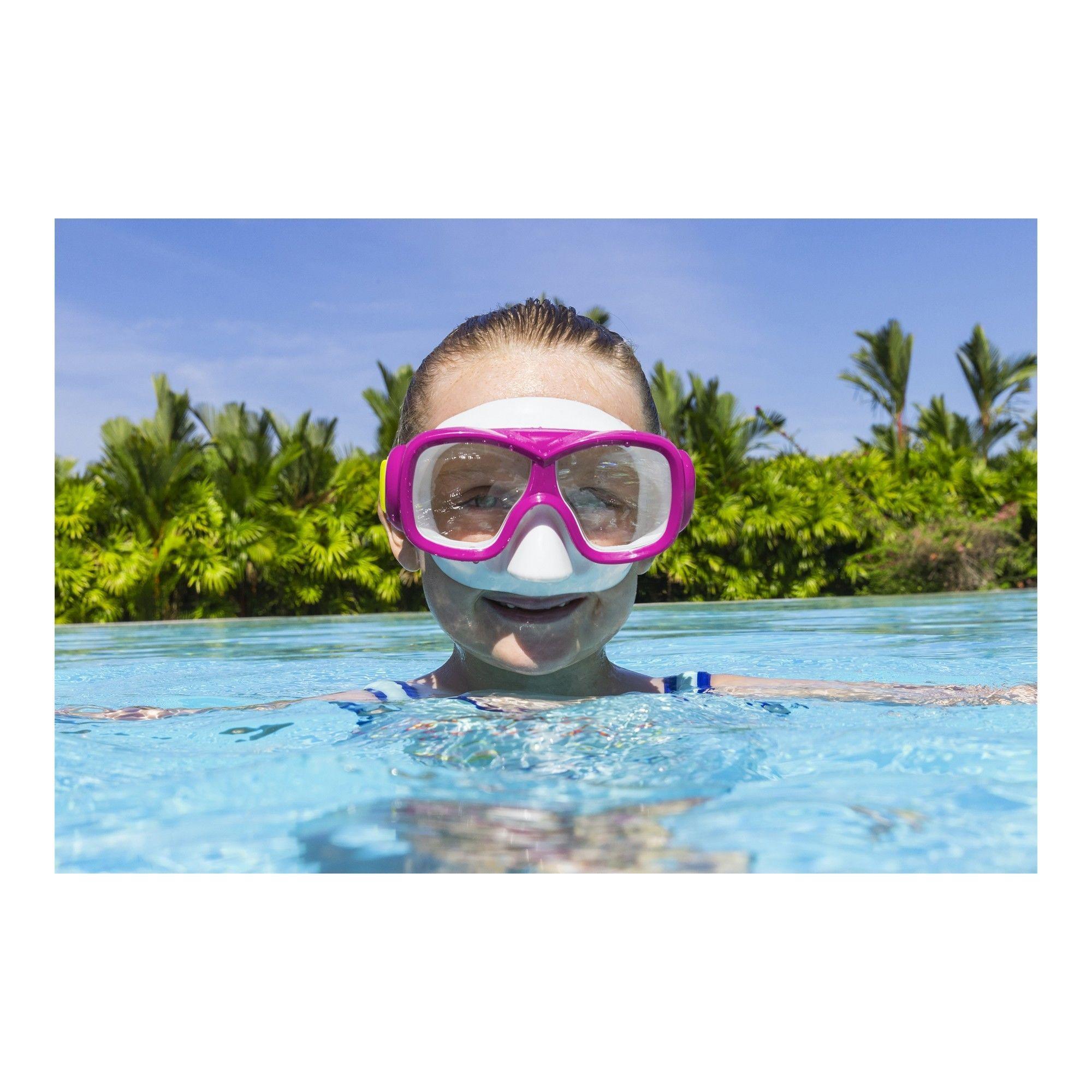 Child wearing pink swim goggles in a pool with clear water, tropical plants in background
