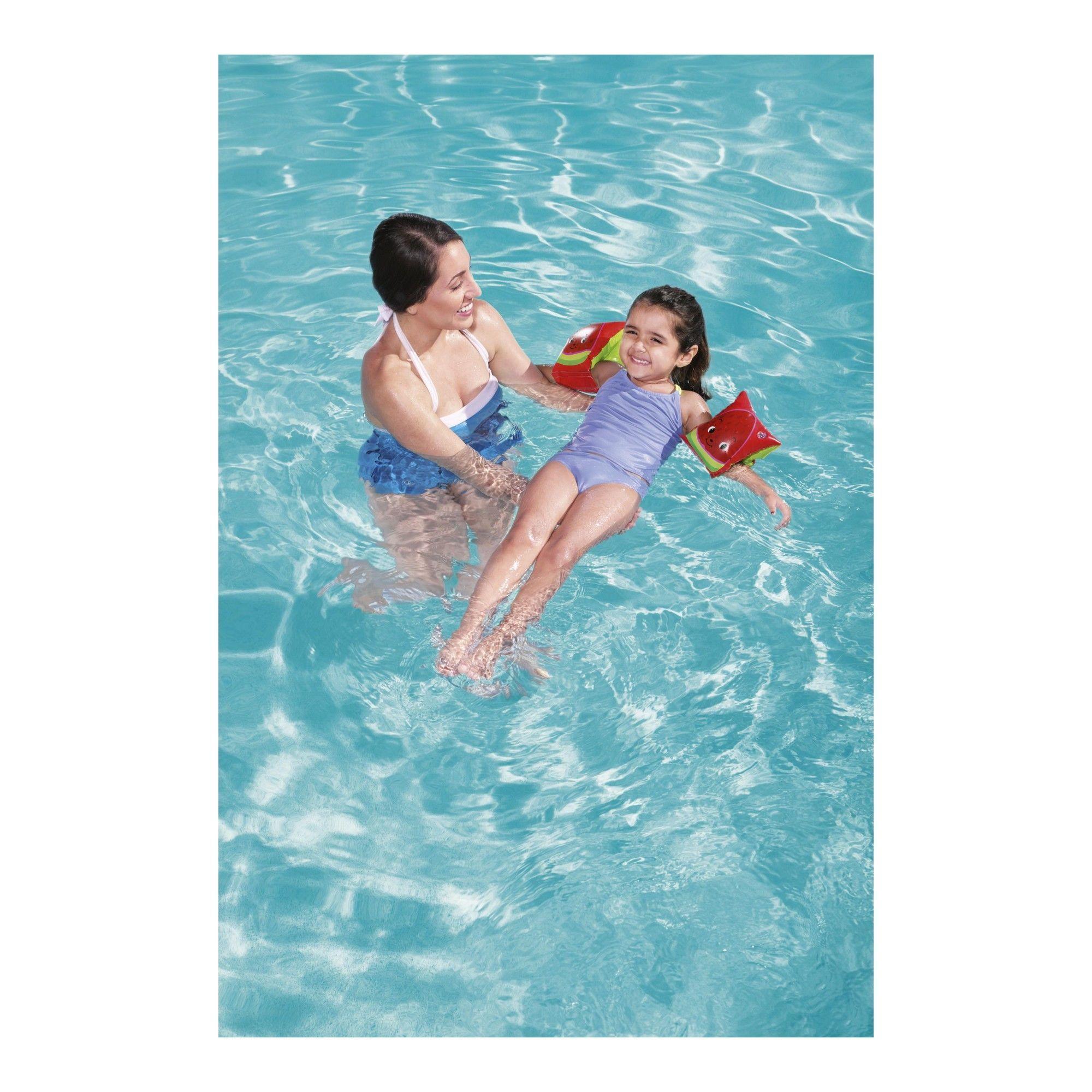 Smiling woman teaching child to swim with red arm floaties in a clear blue swimming pool