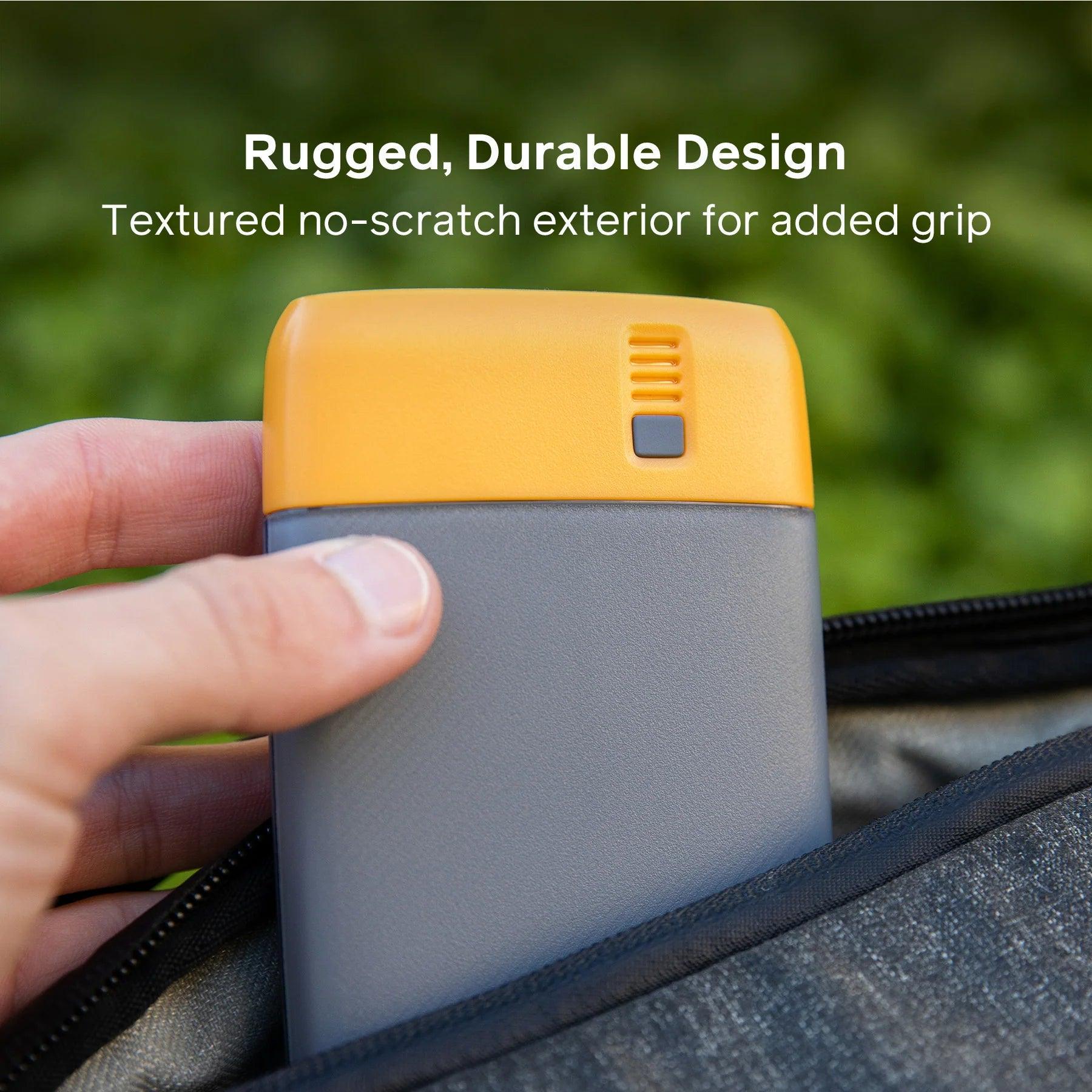 Hand holding a rugged yellow and gray portable power bank outdoors with no-scratch exterior.