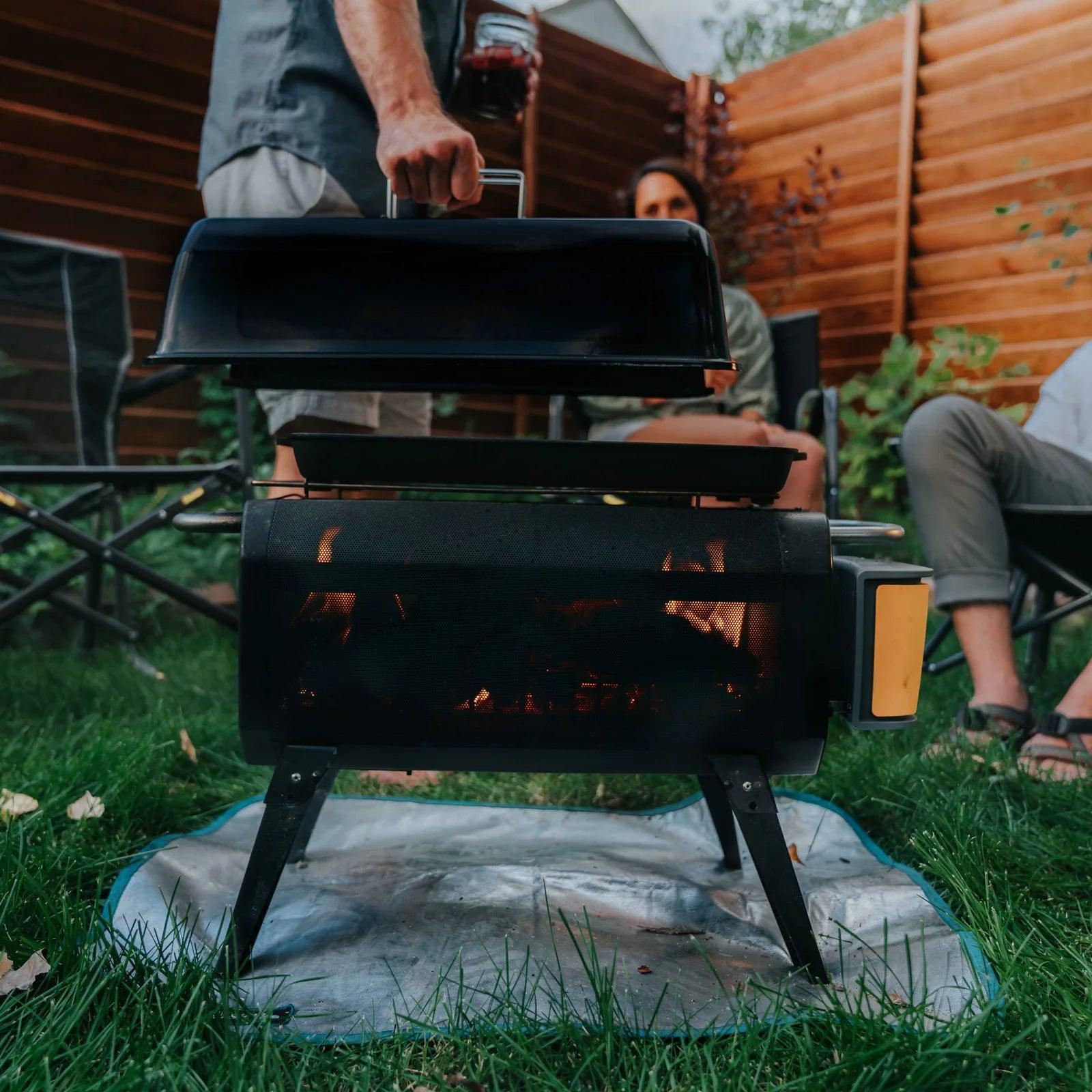 Outdoor BioLite FirePit grill with lid being lifted, people sitting in a backyard