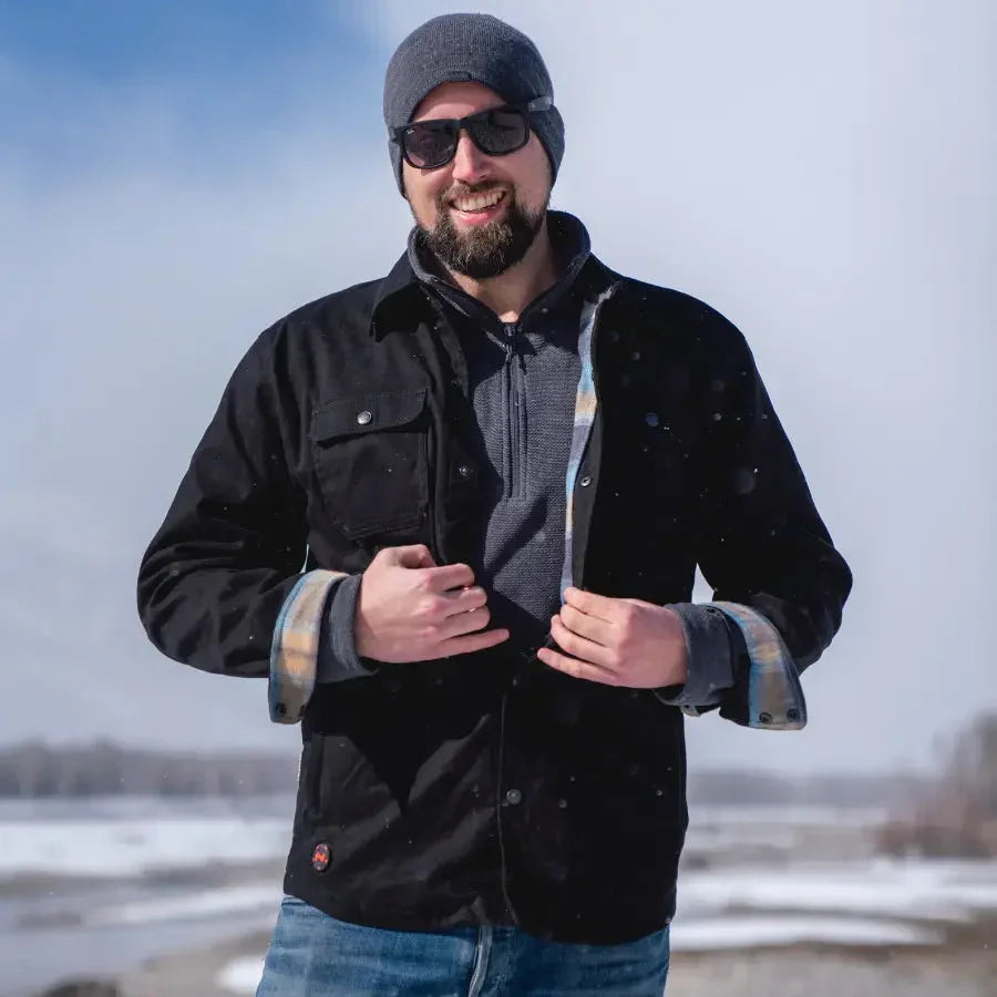 Man in black heated motorcycle jacket and beanie smiling outdoors in snowy landscape
