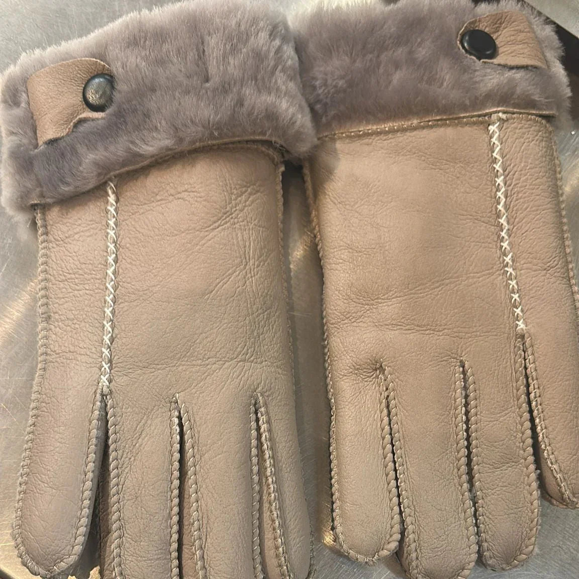 Beige leather gloves with gray faux fur cuffs and decorative stitching on a metallic surface