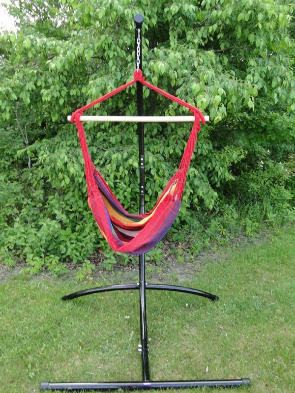 Colorful Brazilian hammock chair with stand set on grass in front of leafy green bushes outdoors.