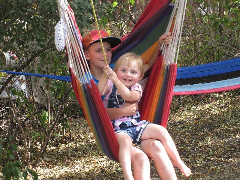 Two children sitting in a colorful Brazilian hammock chair outdoors among trees