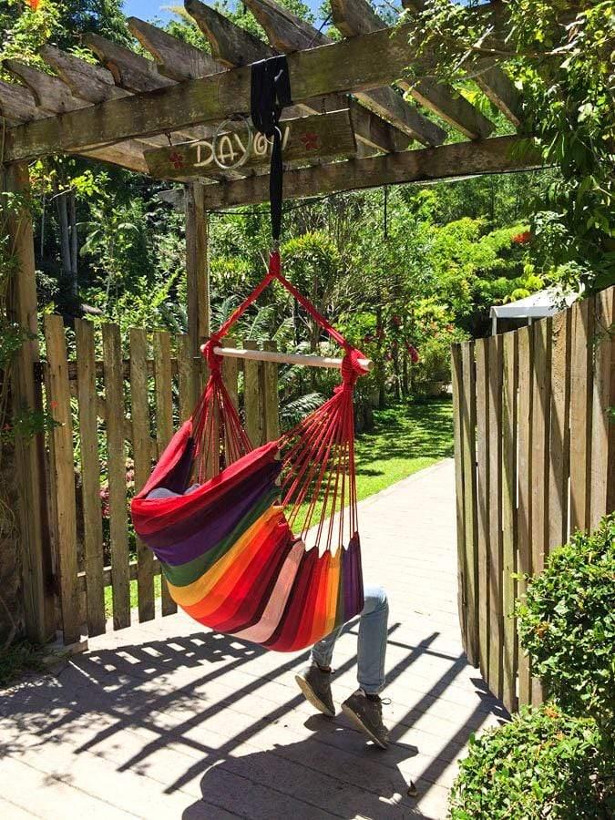 Brazilian hammock chair with rainbow stripes hanging outdoors on a wooden pergola in a garden.