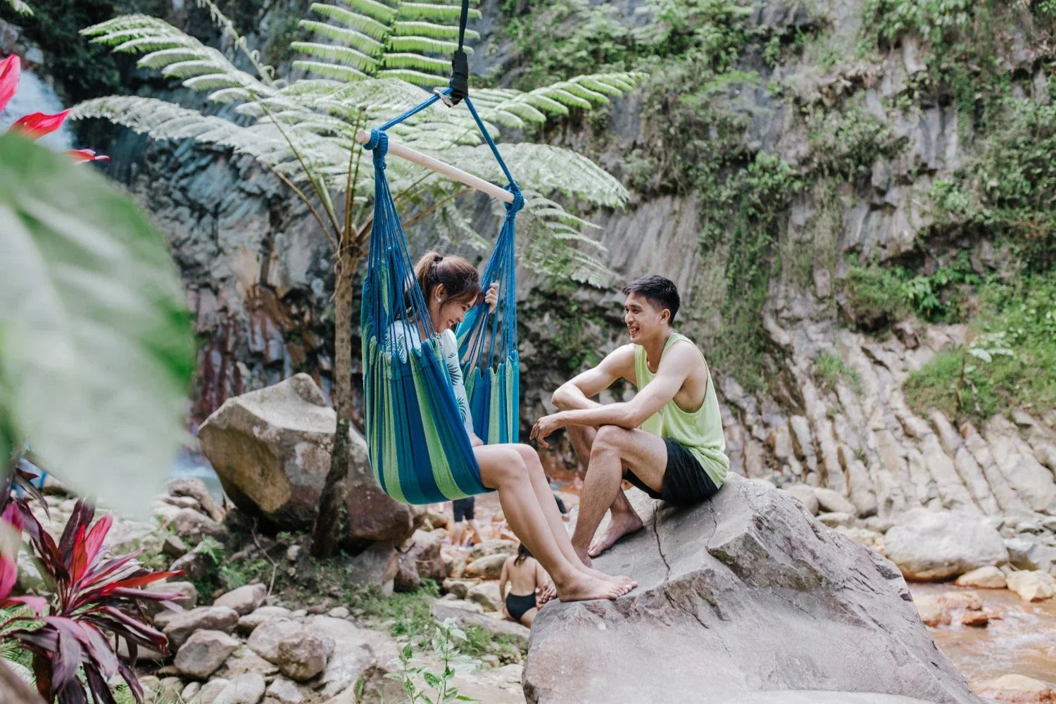 Couple relaxing outdoors, woman in a blue hammock chair, man sitting on rock, lush nature