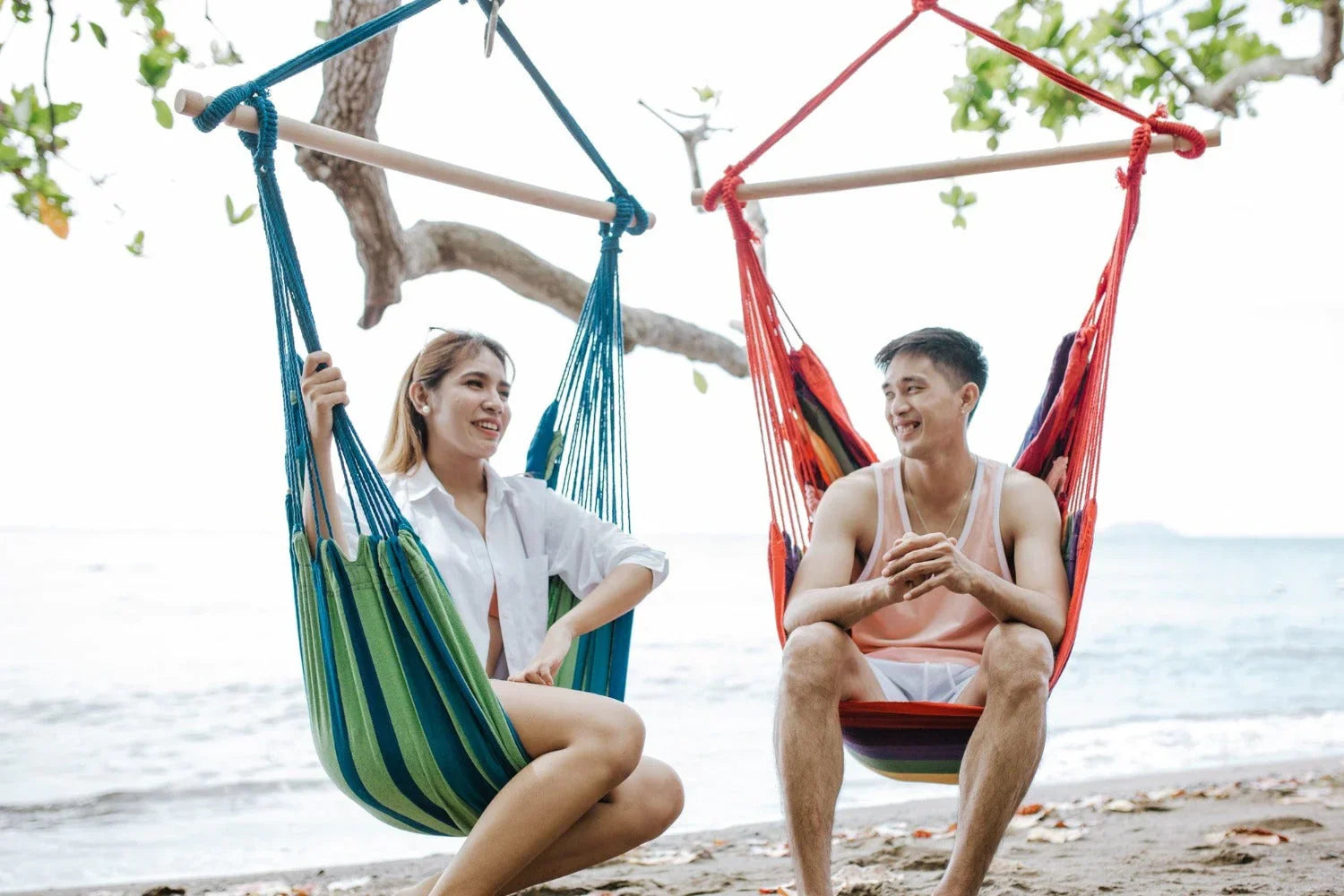 Smiling couple relaxing in colorful hammock chairs by the beach under tree shade