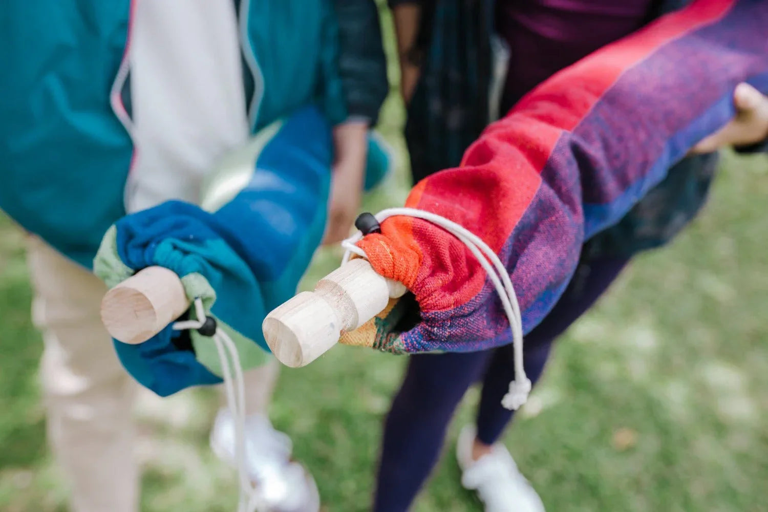 Two people holding colorful fabric bags with wooden handles outdoors on grass
