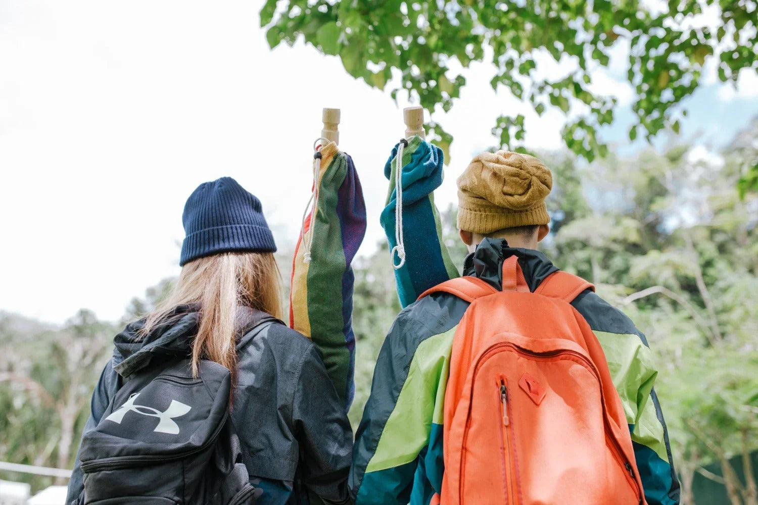 Two kids with backpacks and beanies outdoors carrying rolled-up camping gear