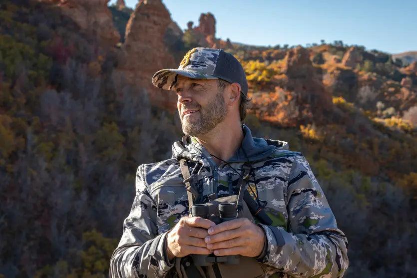 Man in camo jacket and Browning Pahvant Pro cap outdoors with binoculars, rocky background