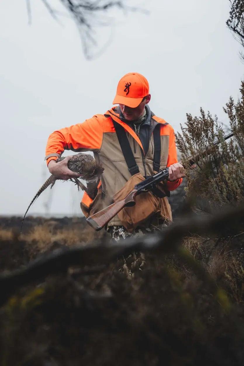 Hunter in upland hunting jacket and orange cap holding pheasant and shotgun in field