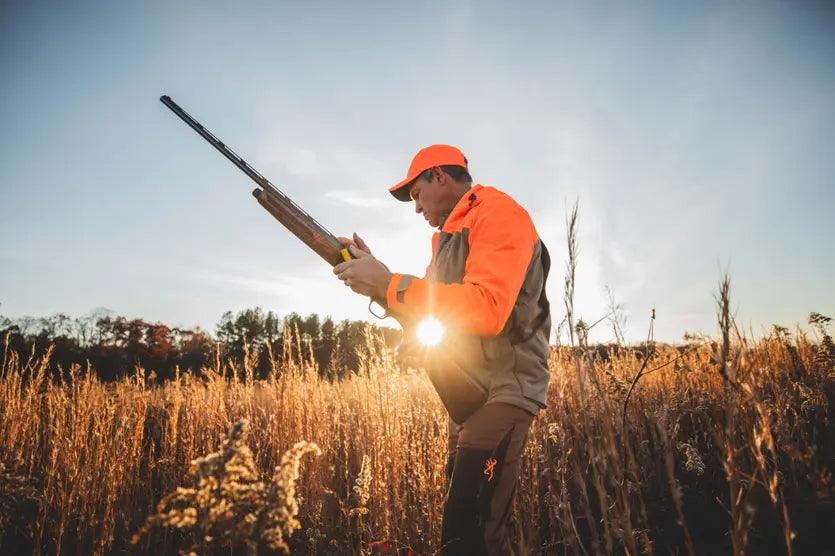 Hunter in orange upland hunting jacket and cap holding shotgun in tall grass field at sunset