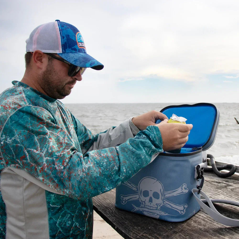 Man in blue fishing shirt and cap using skull logo soft cooler by the ocean