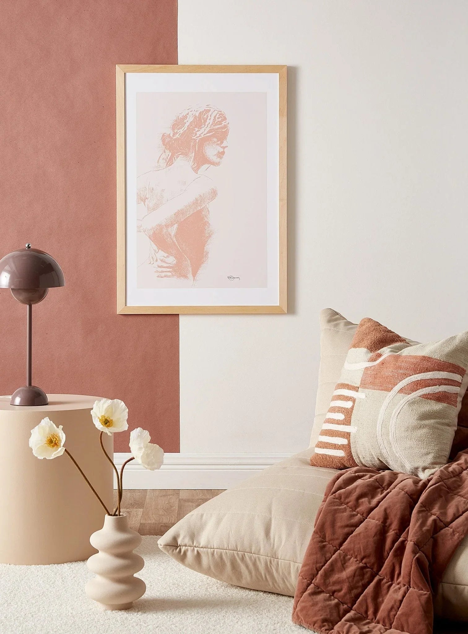 Minimalist living room with beige floor pillow, abstract cushion, flower vase, and framed charcoal woman figure art on a two-tone wall.
