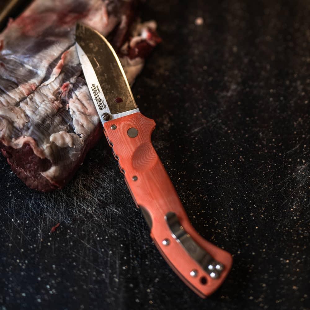 Ultimate Hunter orange folding knife on black surface with raw meat, hunting gear