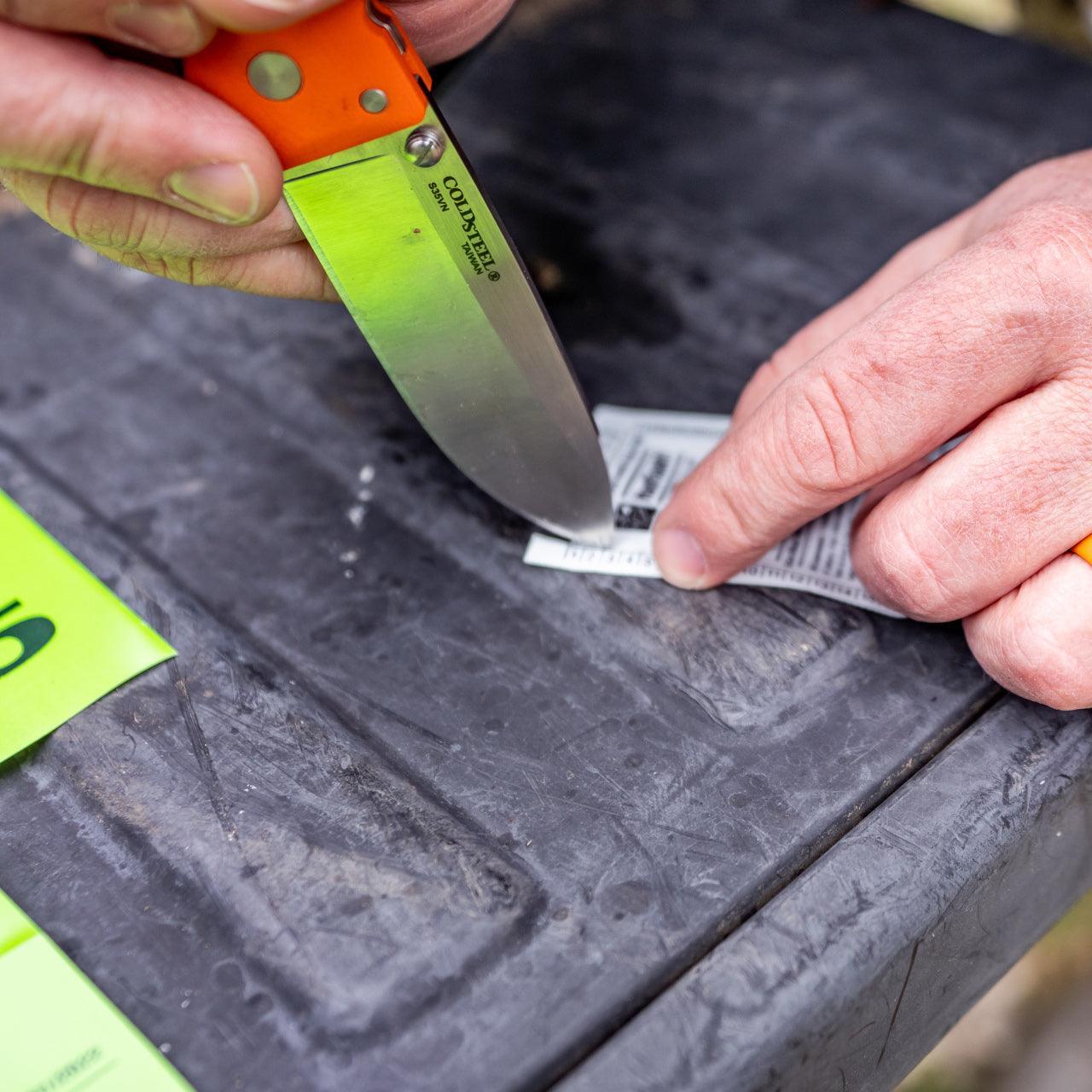 Hand using orange Cold Steel knife to cut paper tag on black outdoor surface