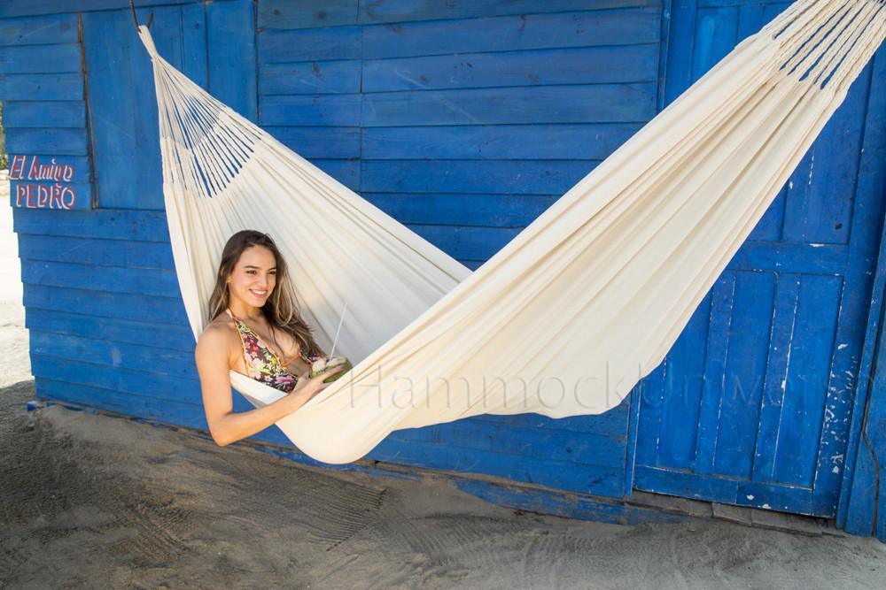 Woman relaxing in a white Colombian hammock in front of a blue wooden wall on sandy ground.