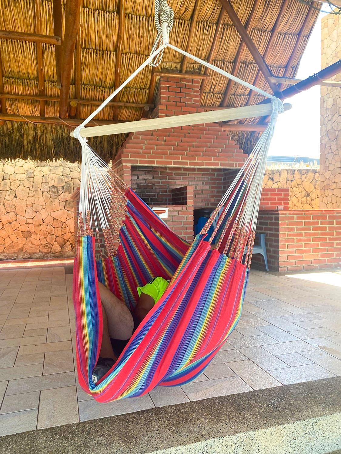 Colorful hammock chair hanging under thatched patio roof near brick grill and stone wall