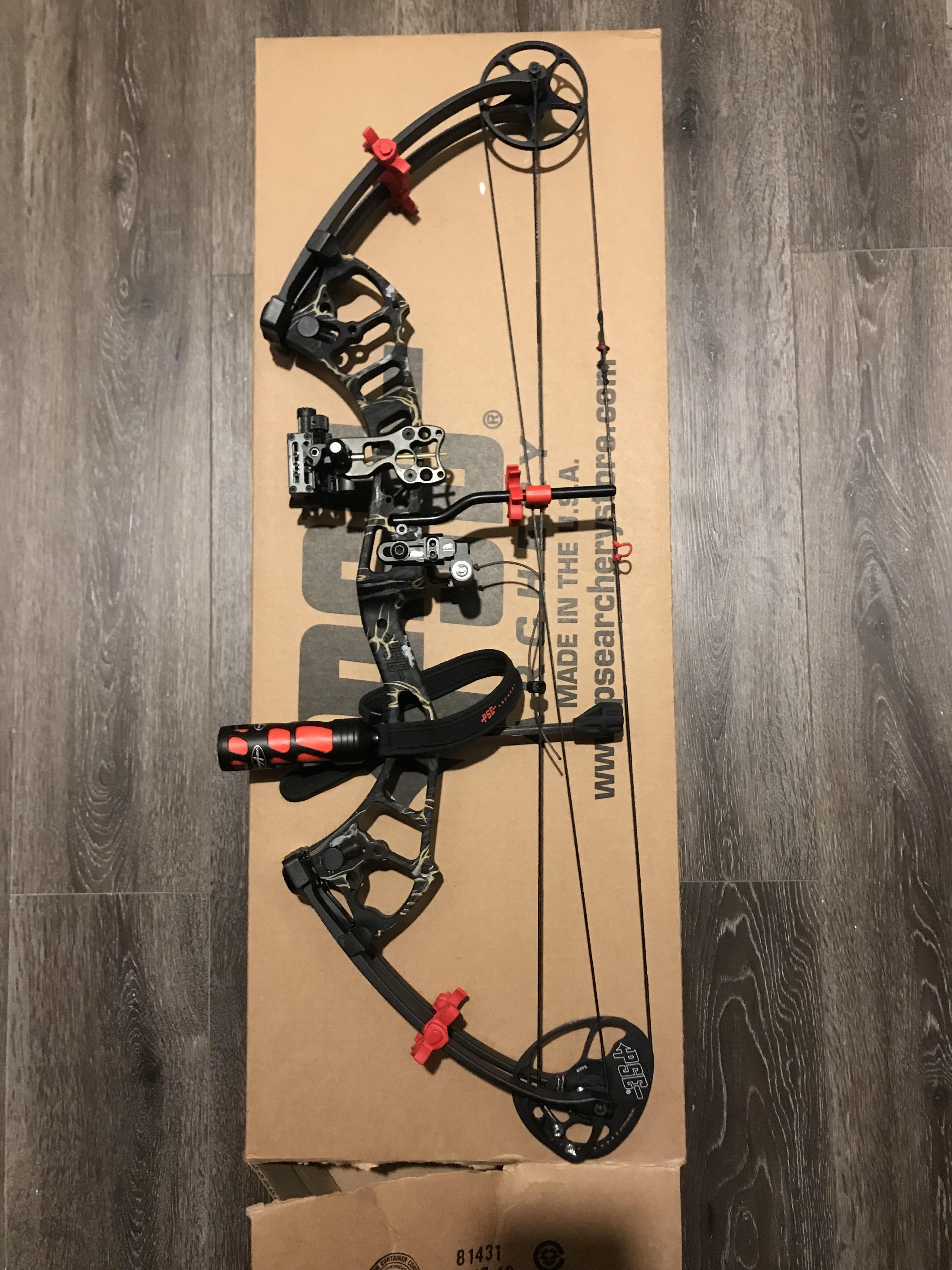 Compound bow with red accents on a cardboard box, displayed on dark wood floor