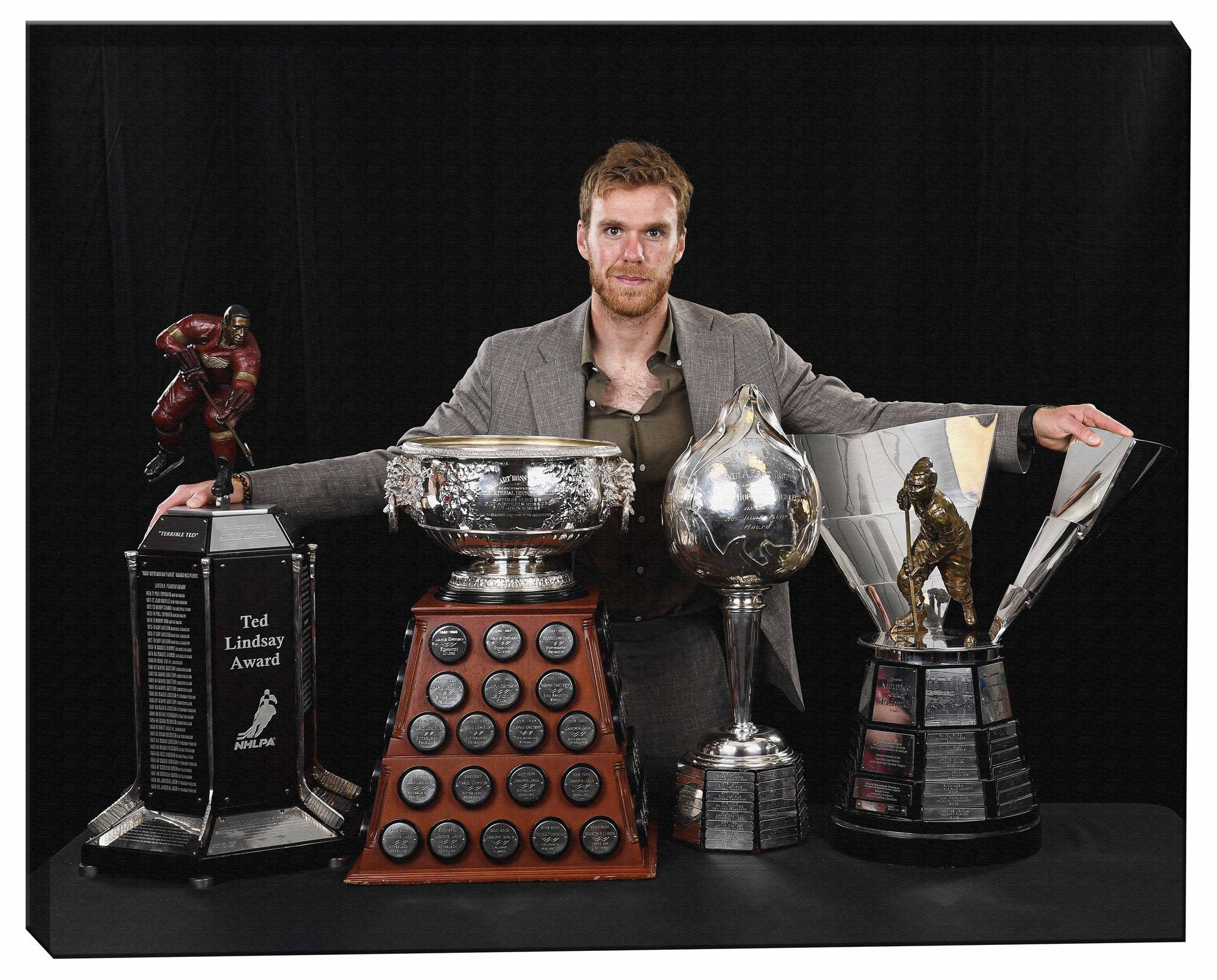 Man in suit displaying NHL hockey trophies including Ted Lindsay Award, Hart Trophy, and Art Ross Trophy against a black background.
