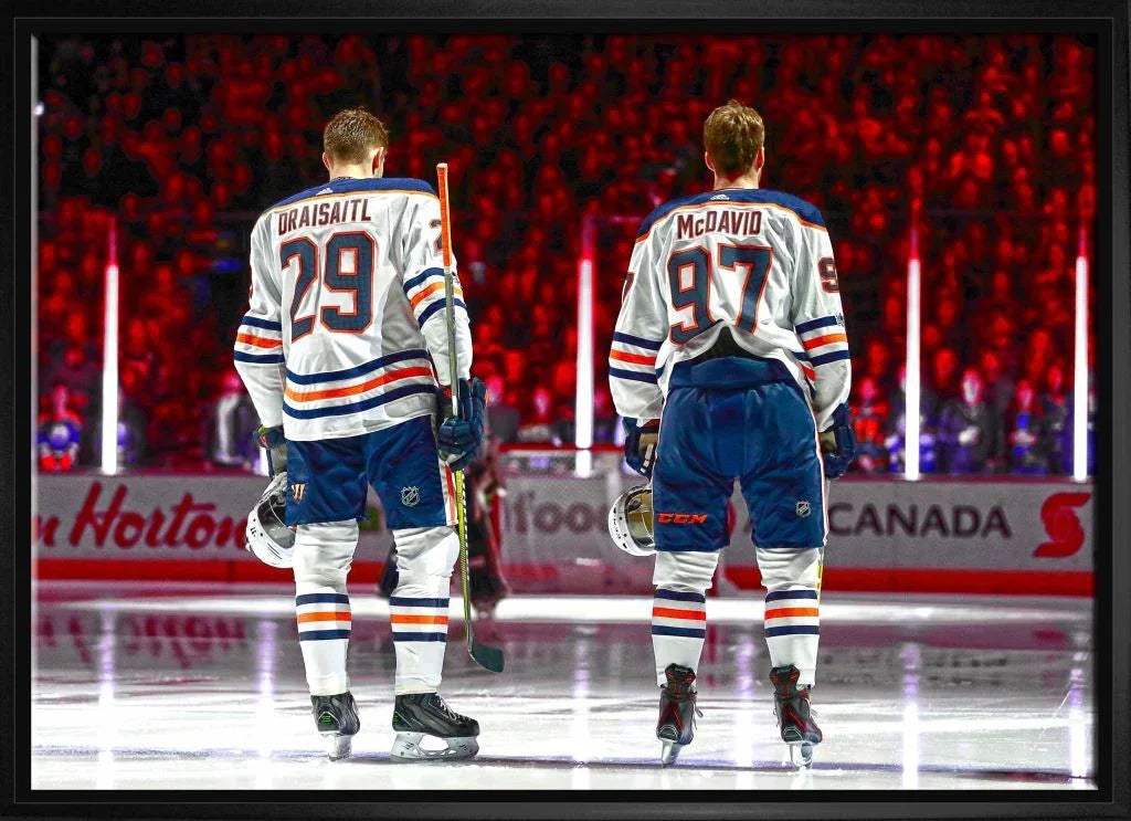 Two Edmonton Oilers hockey players in uniform on ice rink, backs to camera, arena lights.