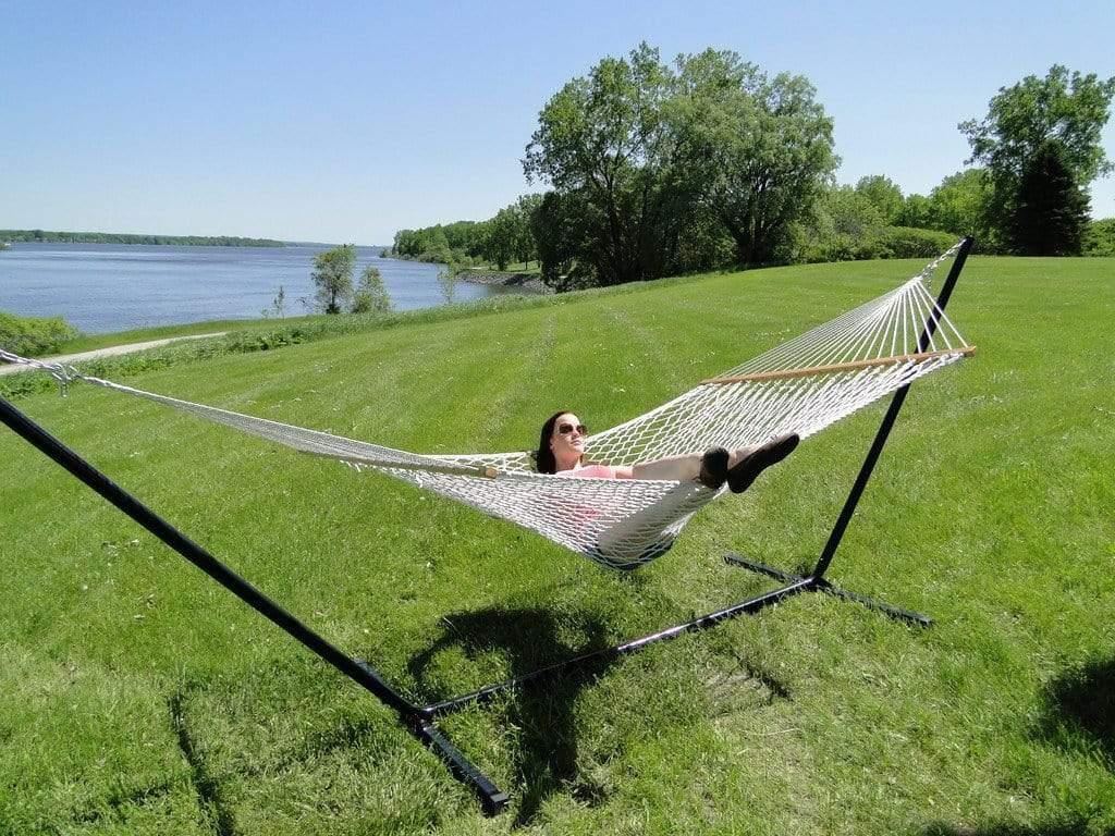 Woman relaxing in cotton rope hammock with tribeam stand on grassy lawn by a lake