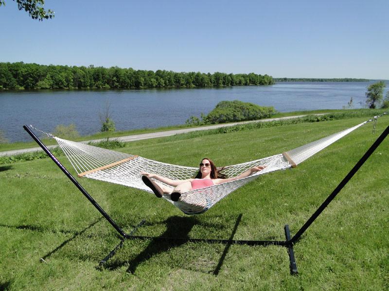 Woman relaxing on a cotton rope hammock with a tri-beam stand by a lake on grassy lawn