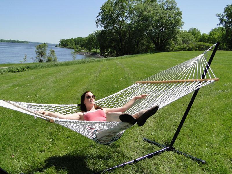 Woman relaxing in a cotton rope hammock on a tri-beam stand by a grassy lakeside