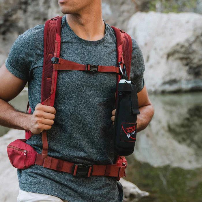 Man hiking outdoors wearing a red backpack with a black holster on the strap near a river.