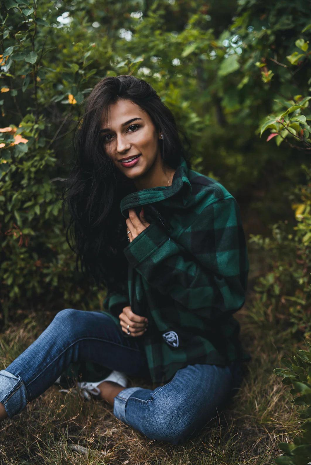 Woman in green plaid fleece jacket and jeans sitting outdoors in a forested area