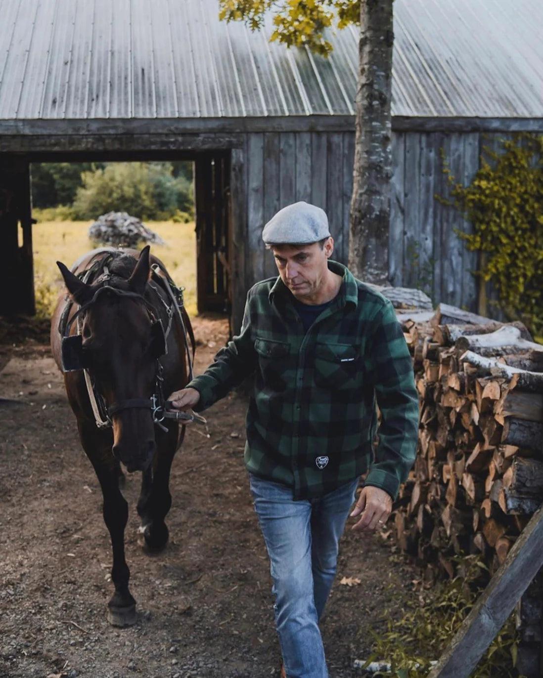 Man in green plaid fleece leading horse by rustic barn and stacked firewood outdoors