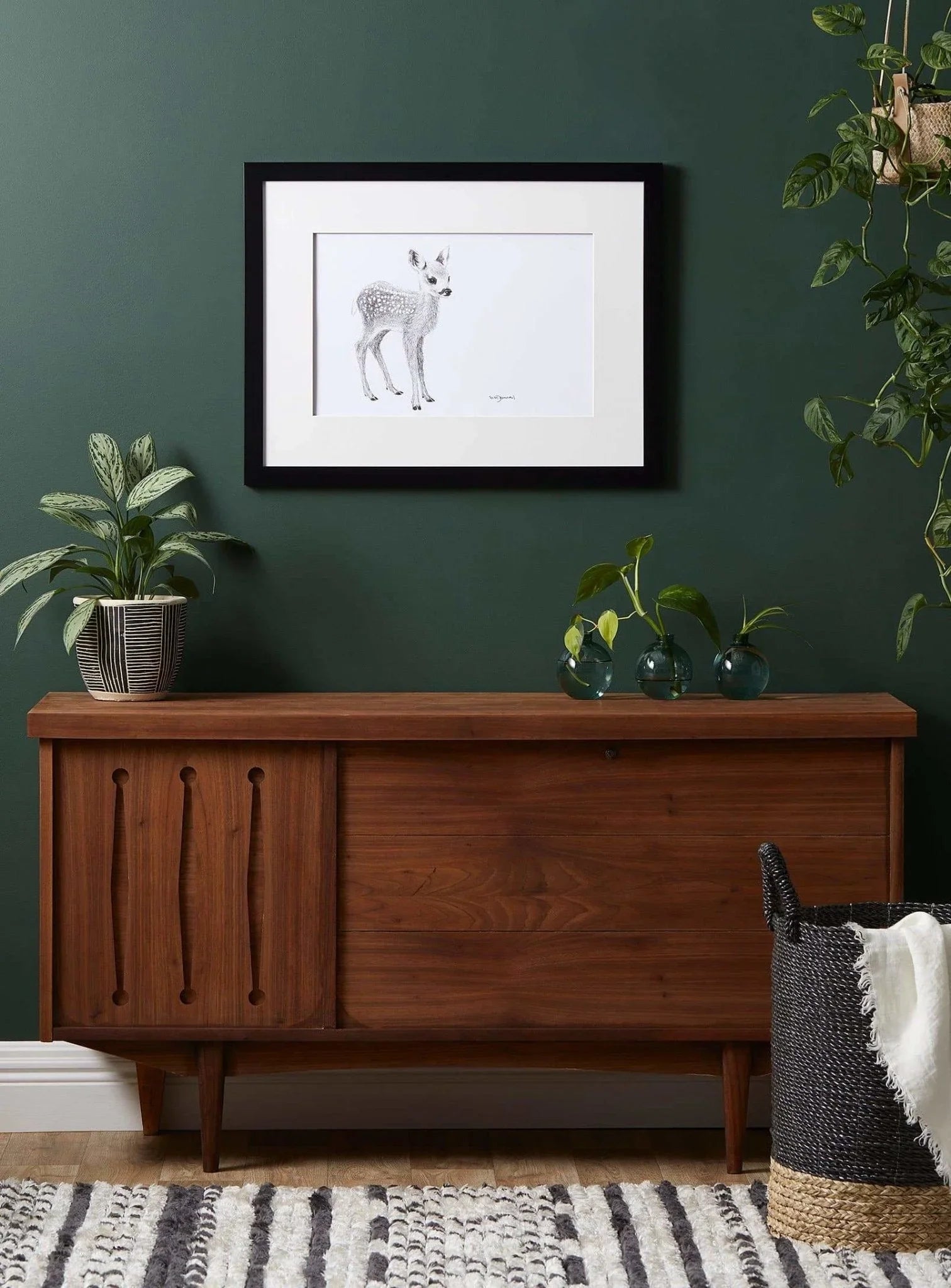 Framed cute baby deer art on green wall above wood sideboard with indoor plants