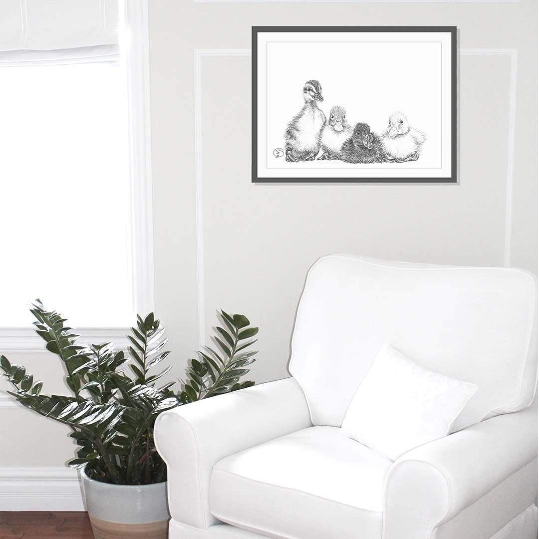 Framed black and white drawing of four baby ducklings hanging above a white armchair.