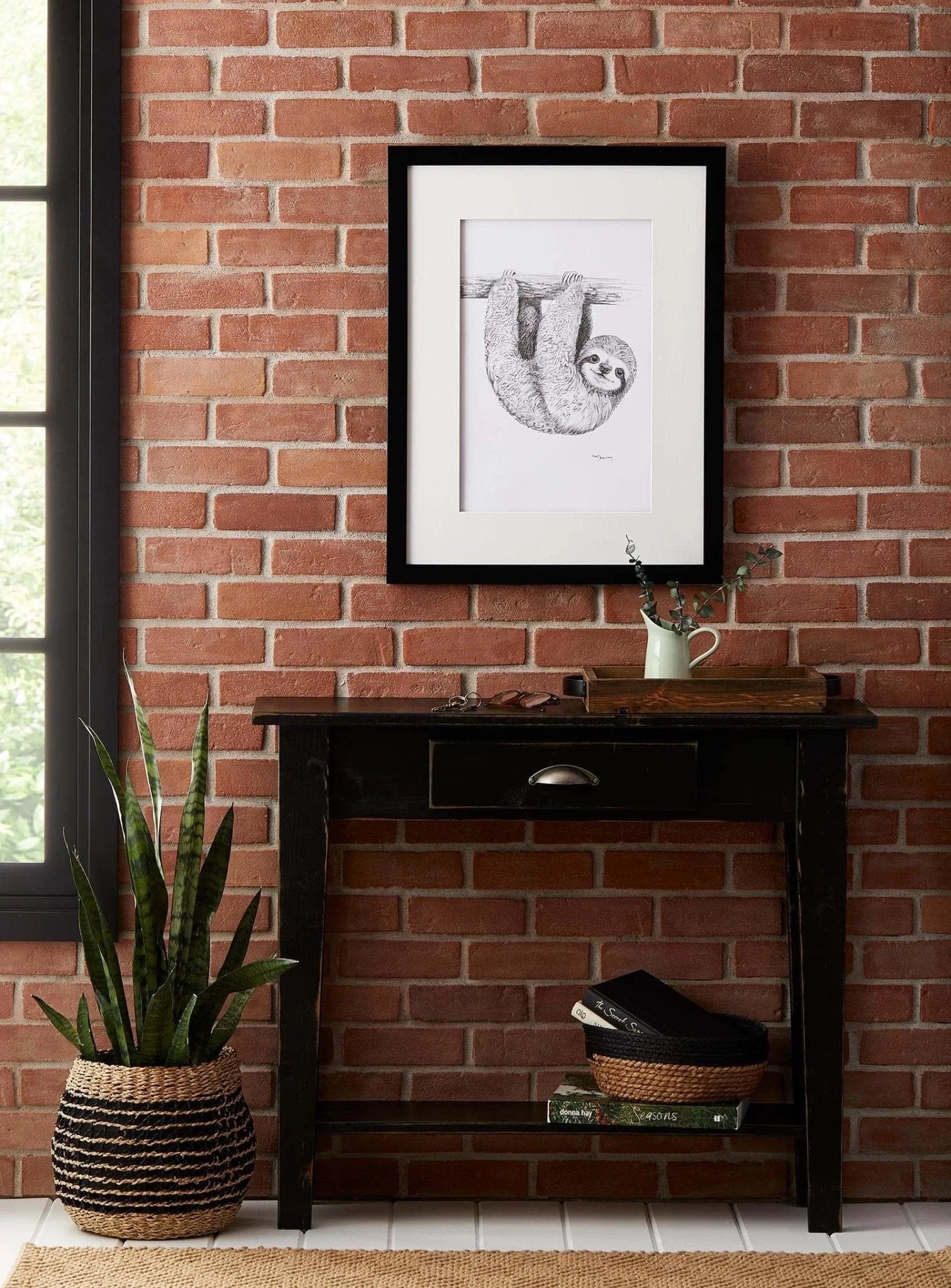 Framed baby sloth drawing on brick wall above black console table with plant decor
