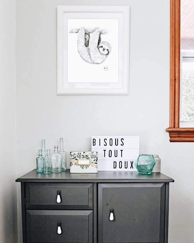 Framed baby sloth drawing on wall above gray cabinet with glass bottles and decor