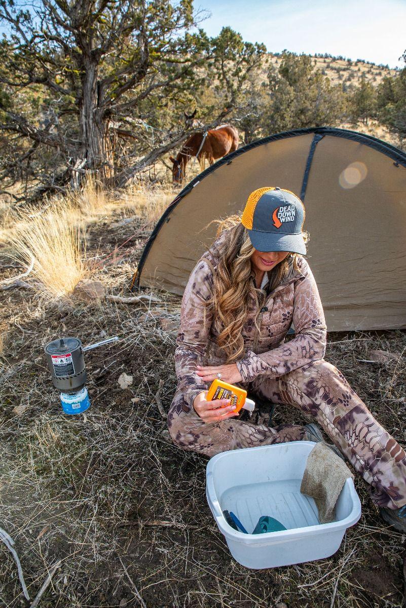 Woman in camo gear at outdoor campsite near tent, holding Dead Down Wind spray, with horse grazing nearby