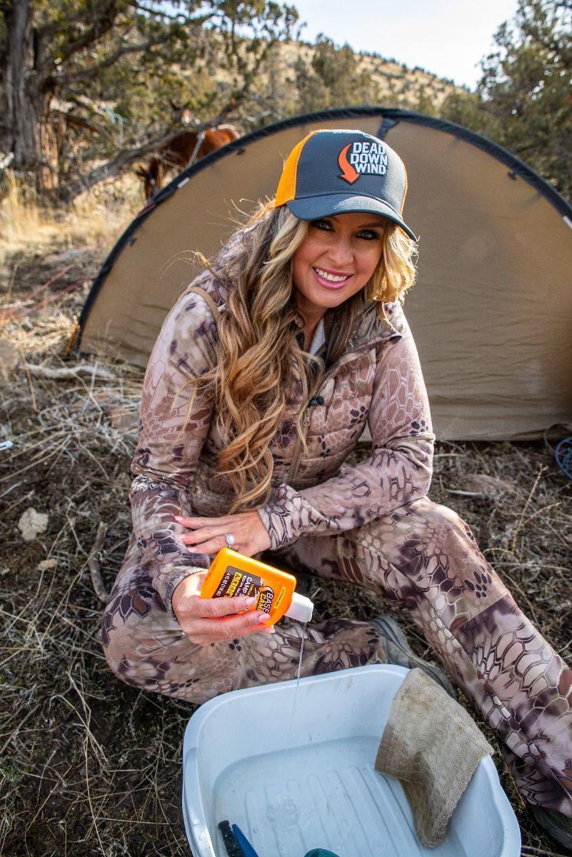 Woman in camo gear camping outdoors, using Dead Down Wind detergent by a tent.