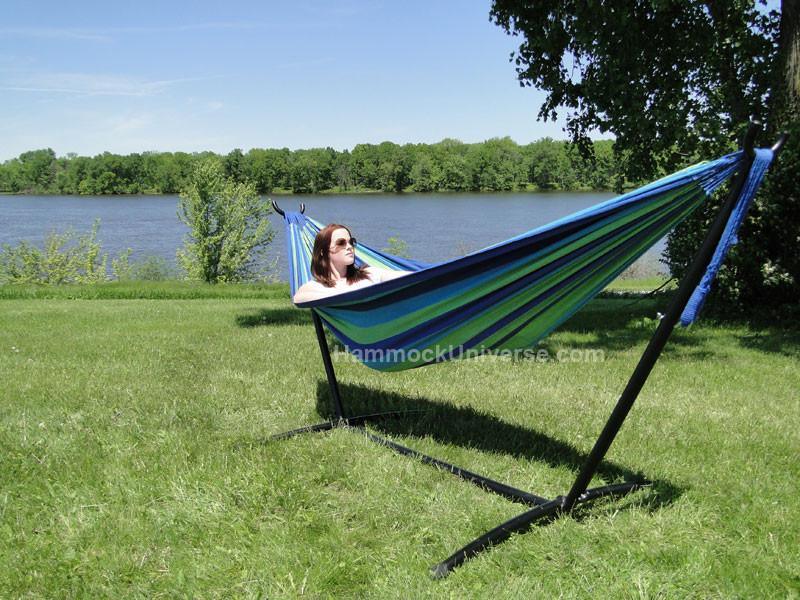 Woman relaxing in a striped Brazilian double hammock on a black stand by a lake in a green park