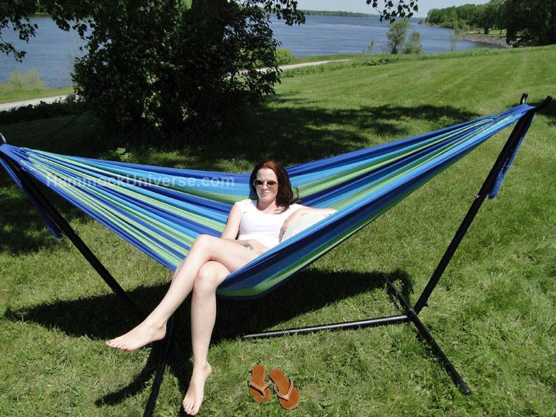 Woman relaxing in a blue and green Brazilian double hammock with stand on grassy lawn by river