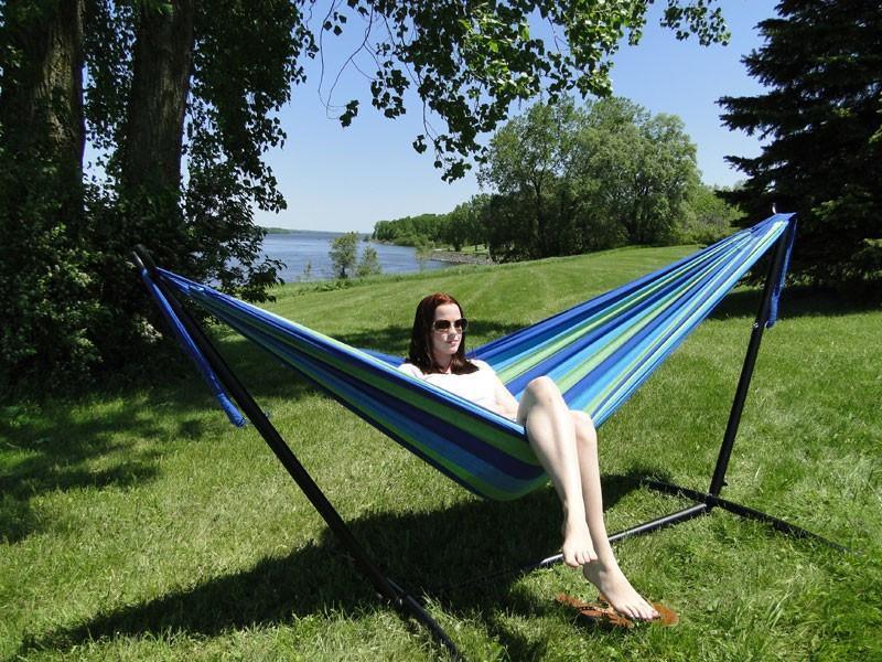 Woman relaxing in blue green Brazilian style double hammock with stand by lake on grassy lawn