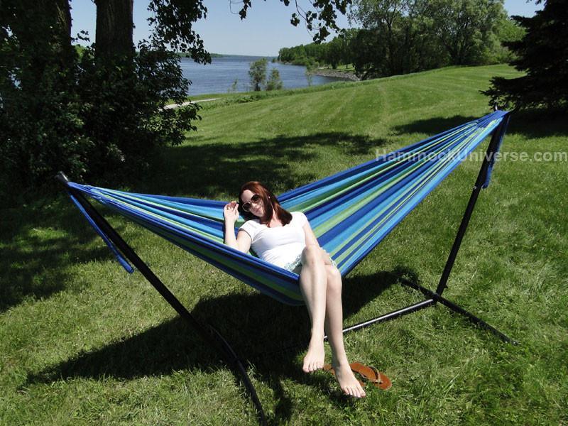 Woman relaxing on a blue striped Brazilian double hammock with stand on a grassy lawn by a lake