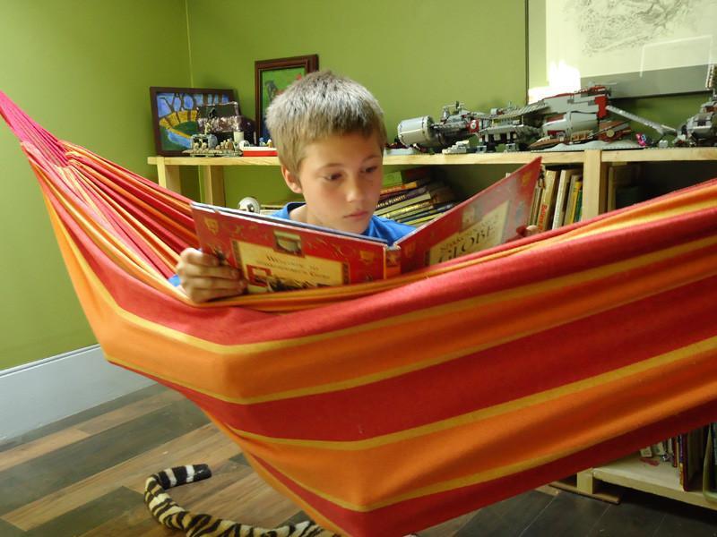 Child reading a book in a red and orange striped Brazilian double hammock indoors