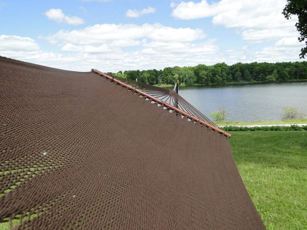 Brown polyester rope hammock with 3-beam stand on grassy lawn near a lake and trees