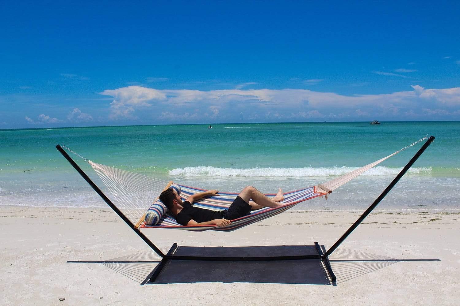 Man relaxing on a striped quilted double hammock with stand at a sunny beach