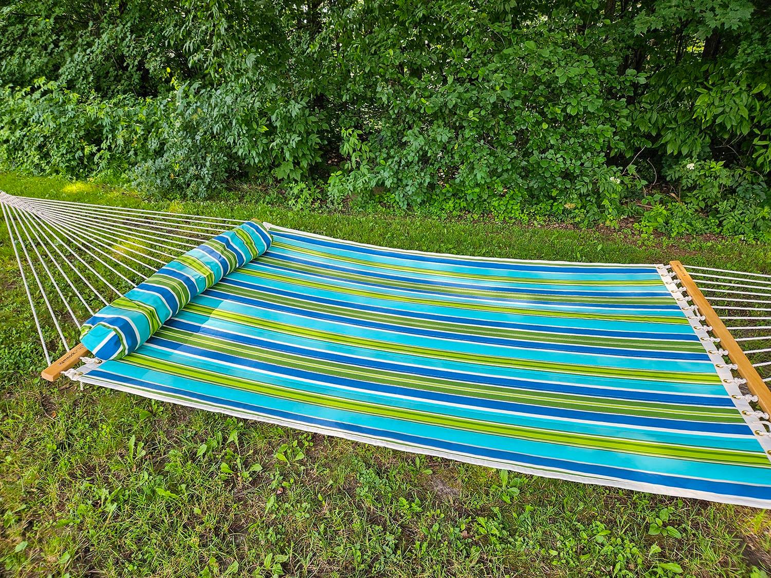 Blue and green striped hammock with pillow on grass by lush green trees outdoors